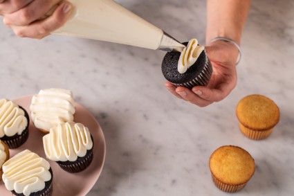 A baker piping a squiggle of frosting onto a cupcake with a pastry bag fitted with a petal tip