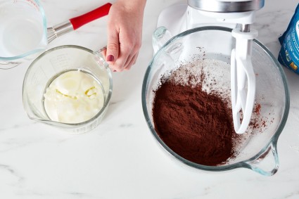 Glass measuring cup of oil and milk next to stand mixer bowl with cocoa powder and other dry ingredients