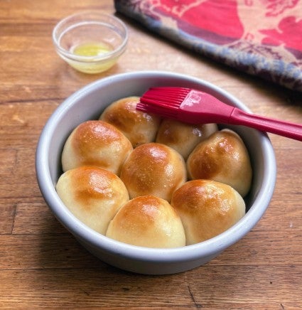 A 6" round pan of golden dinner rolls being brushed with melted butter.