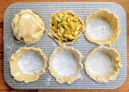 Six mini pie crusts in the wells of a bun pan, some dusted with crust dust, some already filled and topped.