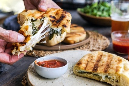 A baker tearing open a freshly baked grilled calzone next to a small bowl of pizza sauce for dipping