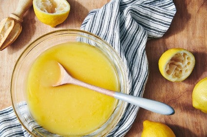 A bowl of homemade lemon curd on a table next to some squeezed lemons