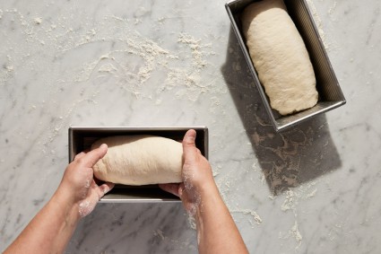 Baker placing shaped sandwich loaf dough into bread pan, next to already filled bread pan