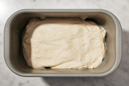 Bread machine bucket with bread dough in it that's been shaped into a loaf