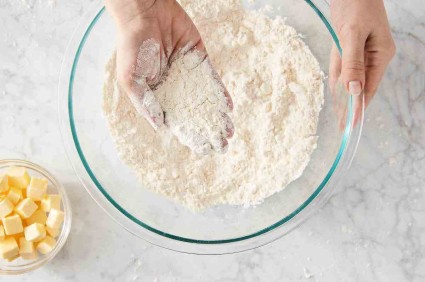 A person's hand holding partially mixed pie crust, showing chunks of butter in flour.