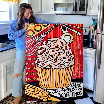 Lacey Hennessey holding up her King Arthur painting, an orange and red depiction of a muffin