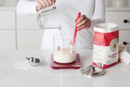 Baker measuring sourdough starter ingredients using glass kitchen scale