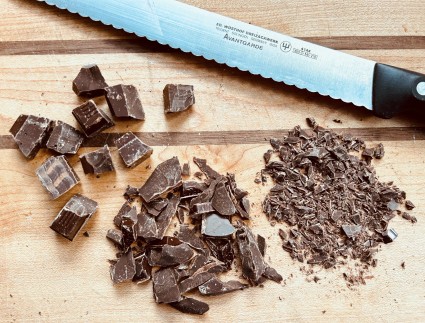 Chunks, shards, and flecks of dark chocolate with a serrated knife on a wooden cutting board. 