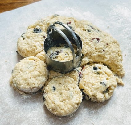 A disc of scone dough on a floured piece of parchment, several round scones cut out of the disc using a sharp metal biscuit cutter.