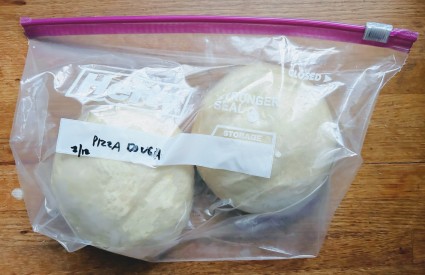 Two balls of pizza dough wrapped in plastic, then sealed in a zip-top plastic bag, ready to go into the freezer for storage.