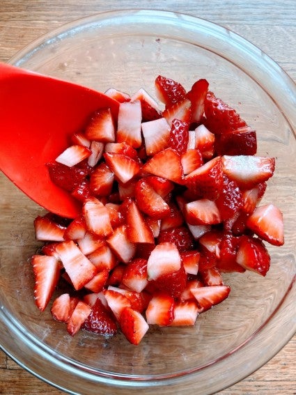 Coarsely chopped strawberries in a glass bowl with a red spoon, glistening with juice.