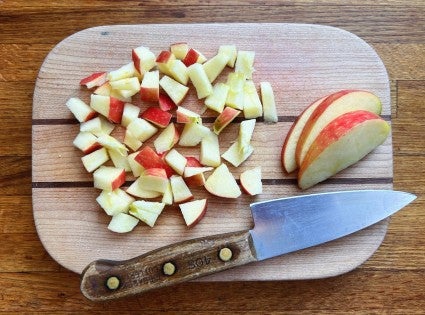 Diced apples, with skin on, on a small wooden cutting board with a knife.