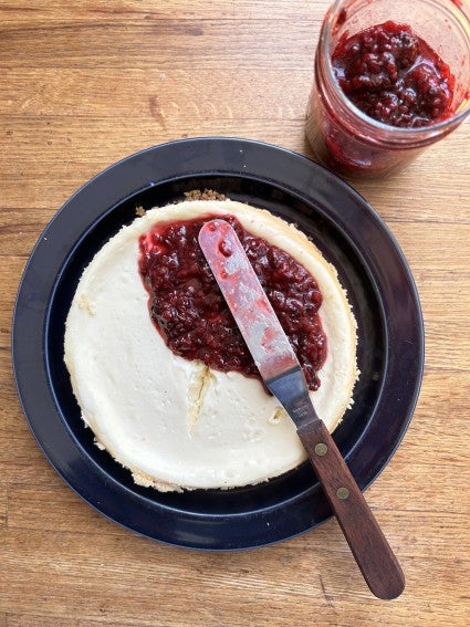 6"cheesecake on a plate, cracked across the center, being spread with raspberry sauce to hid the crack.