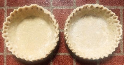 Two pie crusts with tall crimped edges, side by side, ready to go into the oven for prebaking.