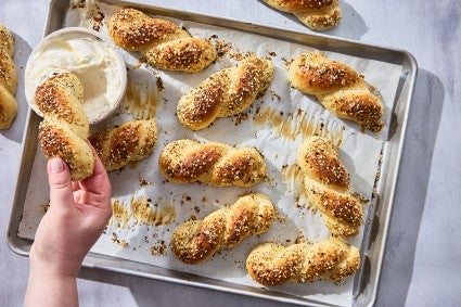 A hand dipping an Everything Bagel Twist into cream cheese along with other bagel twists spread out on a sheet tray and parchment paper.