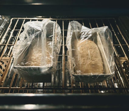 Two loaf pans holding rising dough, covered with plastic see-through shower caps and set on the middle rack of a gently warmed oven.
