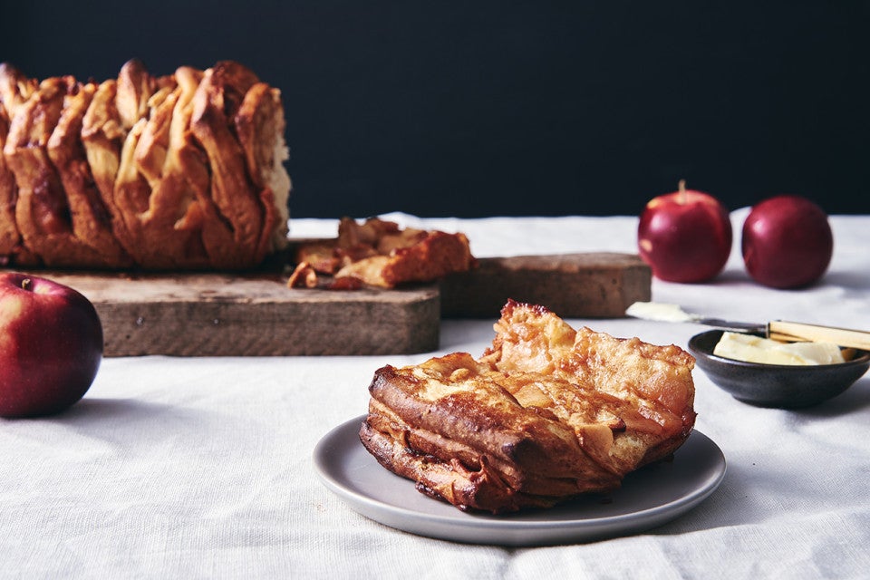 A loaf of Cinnamon Apple Pull-Apart Bread next to a few apples and slice of pull-apart bread on a plate, ready to be eaten.