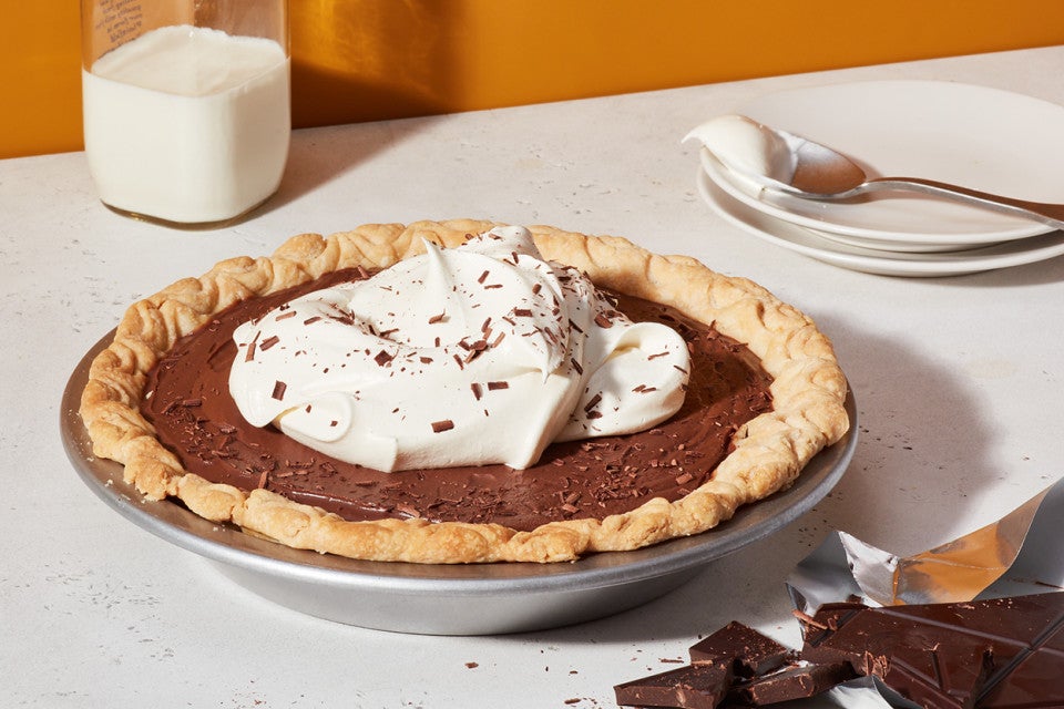 A Chocolate Cream Pie topped with whipped cream next to a stack of plates and a glass of milk