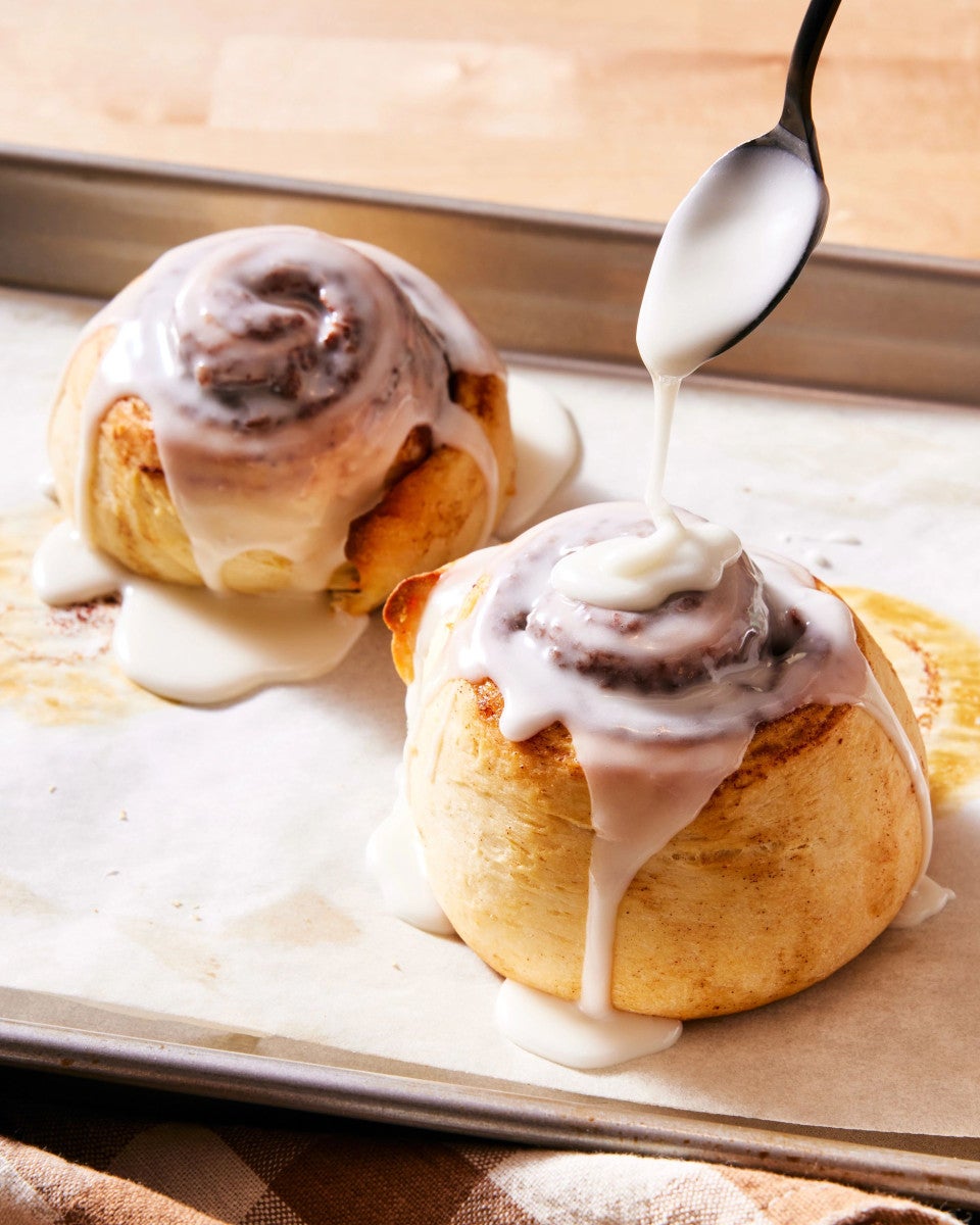 A small batch of two cinnamon rolls on a quarter sheet pan being frosted with icing
