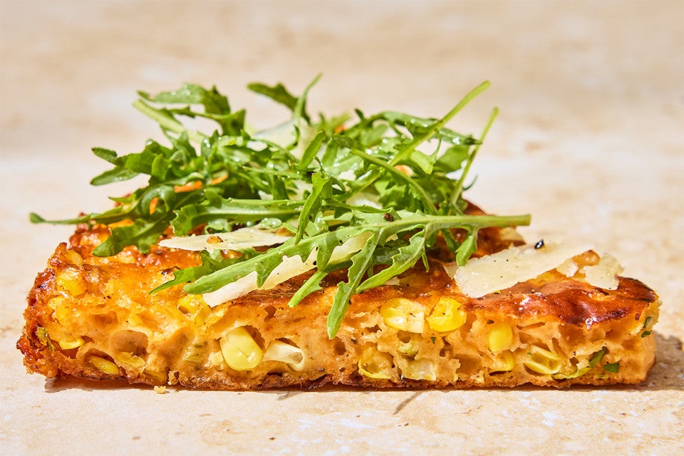 A slice of Giant Cheesy Corn Fritter shown from the side; topped with greens