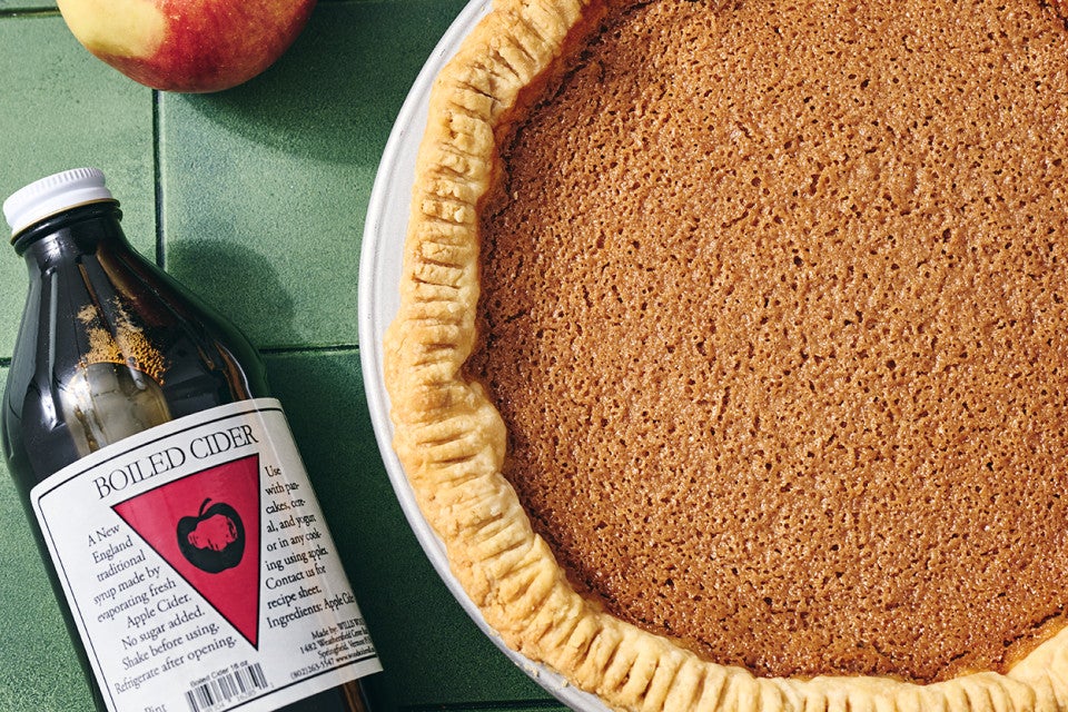 An apple pie seen from above with a smooth texture and a bottle of boiled cider laying on the table next to it.
