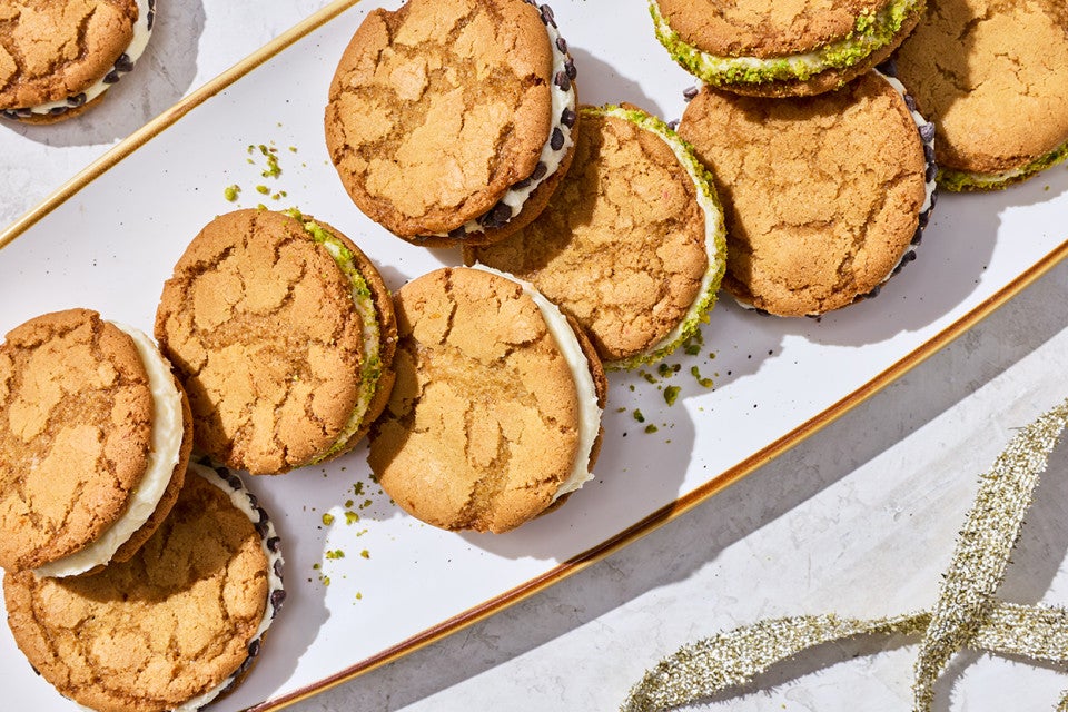 Looking down on a festive plate of cannoli sandwich cookies with cracked tops and crumbs of pistachios.
