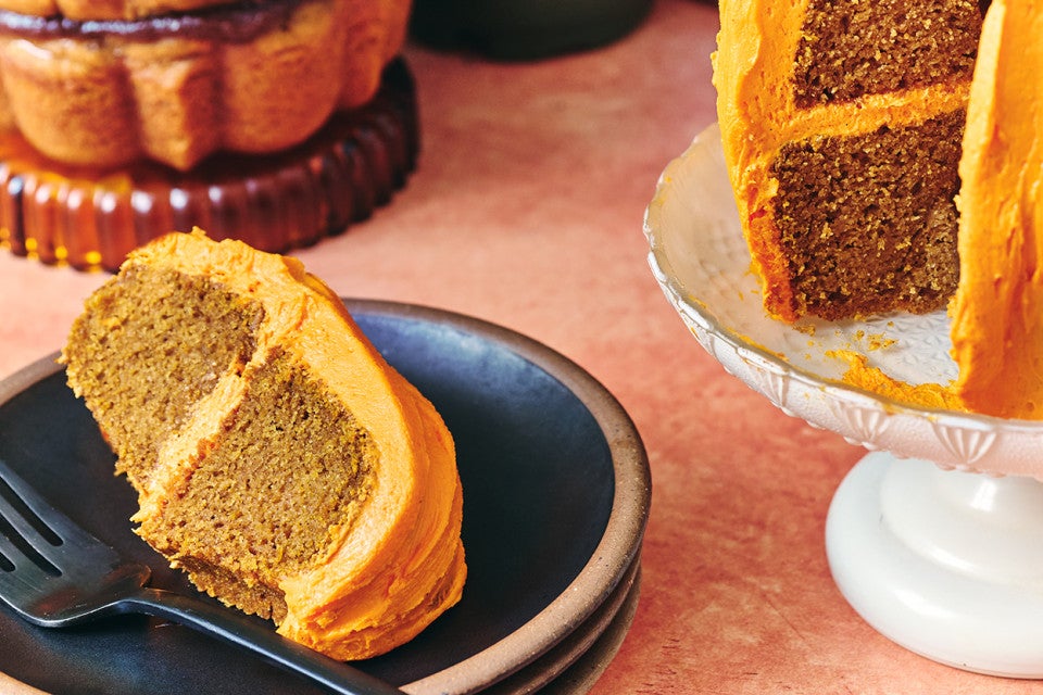 A slice of pumpkin bundt cake on a dark colored plate layered and covered with orange frosting.