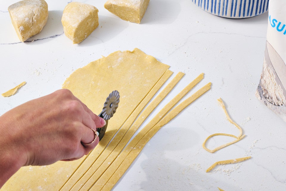 Photograph of a person using a rolling cutter to create gluten free pasta strands from a sheet of raw dough.