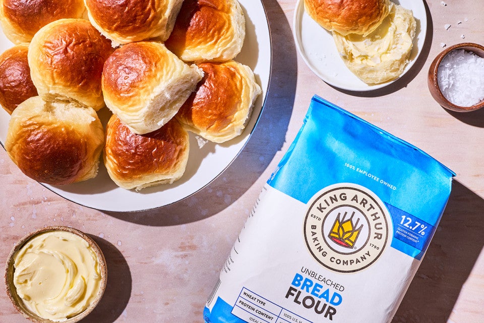 A plate with browned dinner rolls, a dish with butter, another with a sliced roll, and a bag of King Arthur Bread Flour.