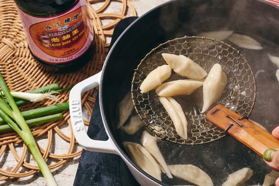 A handheld noodle mesh scoop picking up hand cut noodles from a pot of boiling water seen from above the food.