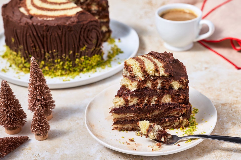 Showing Souche de Noël sliced on a plate with layers (rings) of vanilla and chocolate cake and little brown Christmas trees on a counter with the rest of the cake in the background.