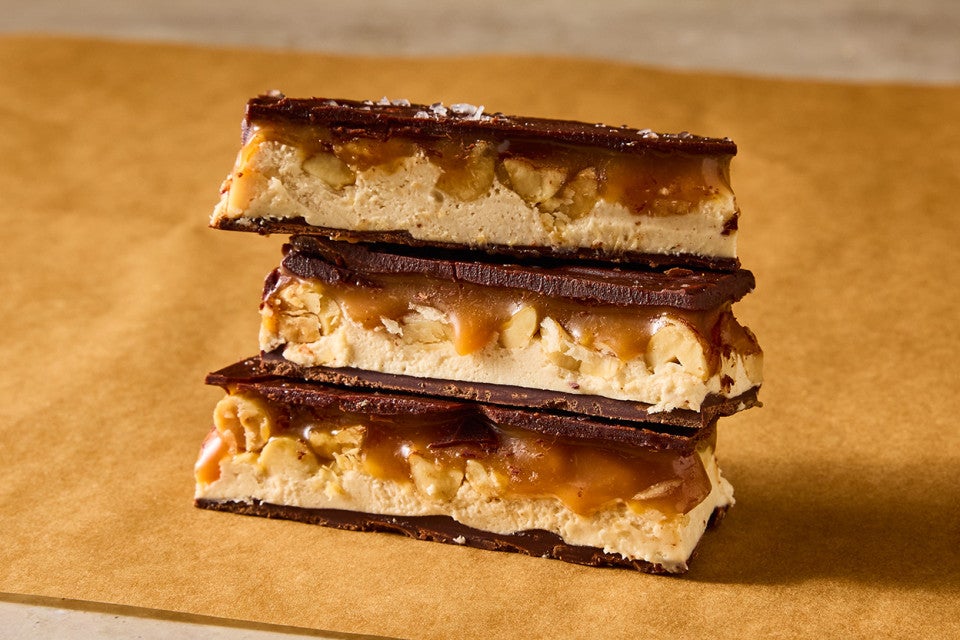 Three sliced chocolate peanut nougat bars stacked on brown parchment and showing the layers of chocolate, nougat, caramel, and peanuts.