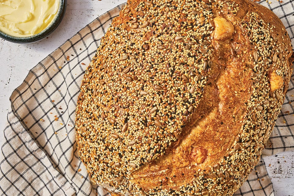 A loaf of Parmesan Black Pepper Sourdough Bread shown on a gingham kitchen towel coated in seeds and flavorful bread topping.
