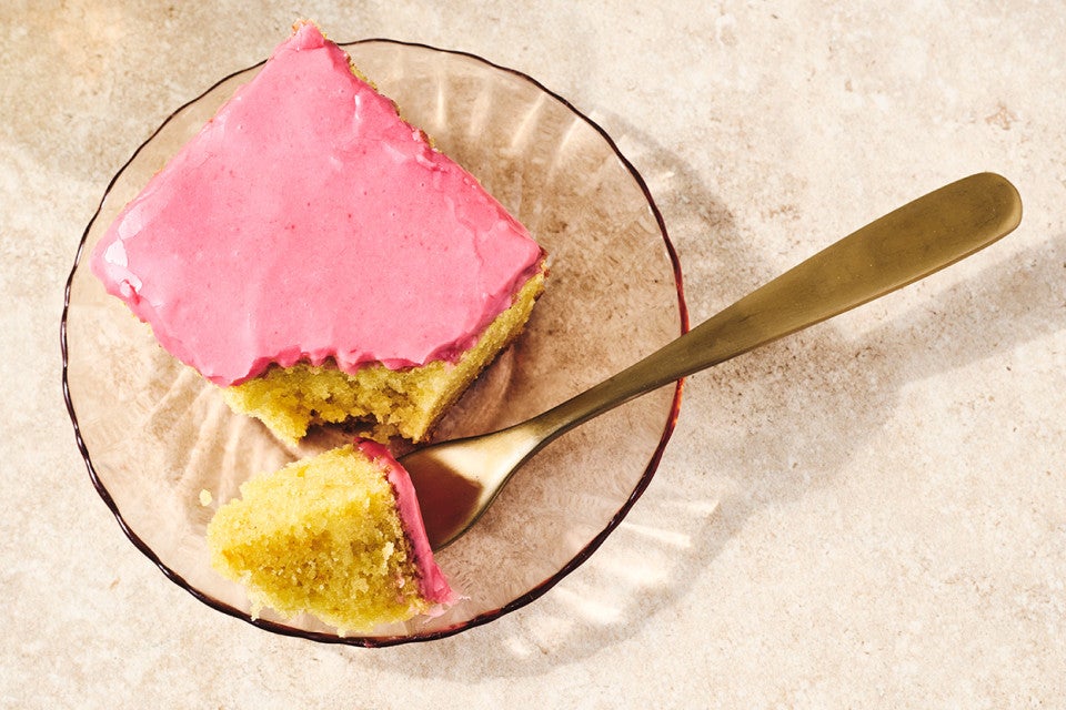 A slice of Cranberry-Orange Olive Oil Cake on a glass plate with a bite on a fork showing a moist golden cake and pastel pink frosting.