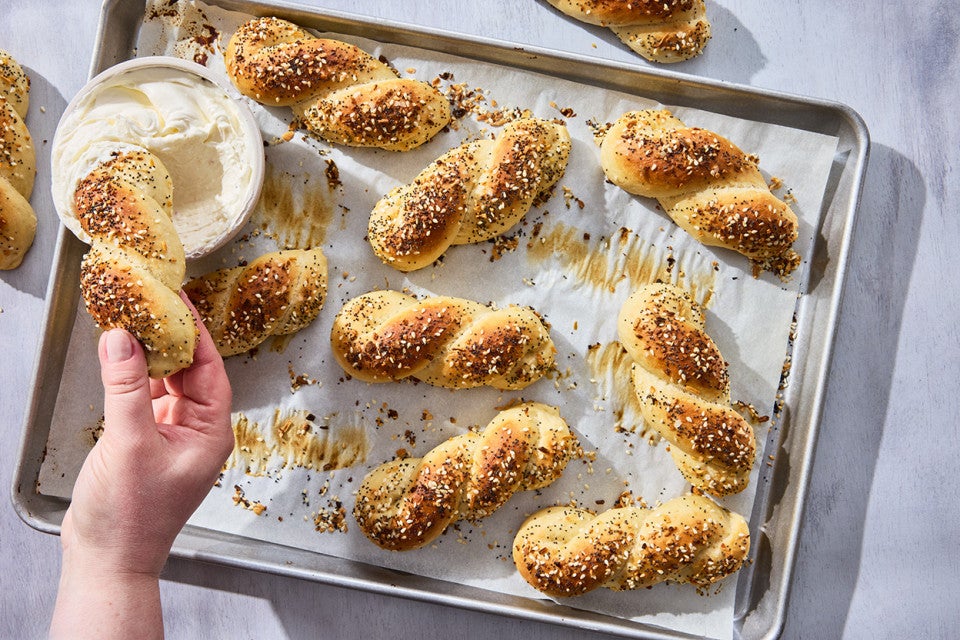 A hand dipping an Everything Bagel Twist into cream cheese along with other bagel twists spread out on a sheet tray and parchment paper.