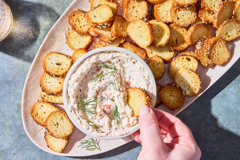 A hand dipping an everything bagel chip into Salmon Cream Cheese dip.