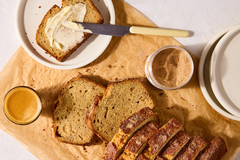A sliced loaf of High-Altitude Banana Bread laid out on a parchment with a buttered slice on a plate, a cup of coffee, and cinnamon sugar in a dish.