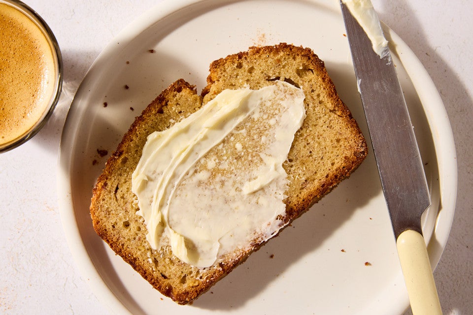 A slice of High-Altitude Banana Bread buttered and laying on a light plate with a butter knife laid on it's side next to the bread.