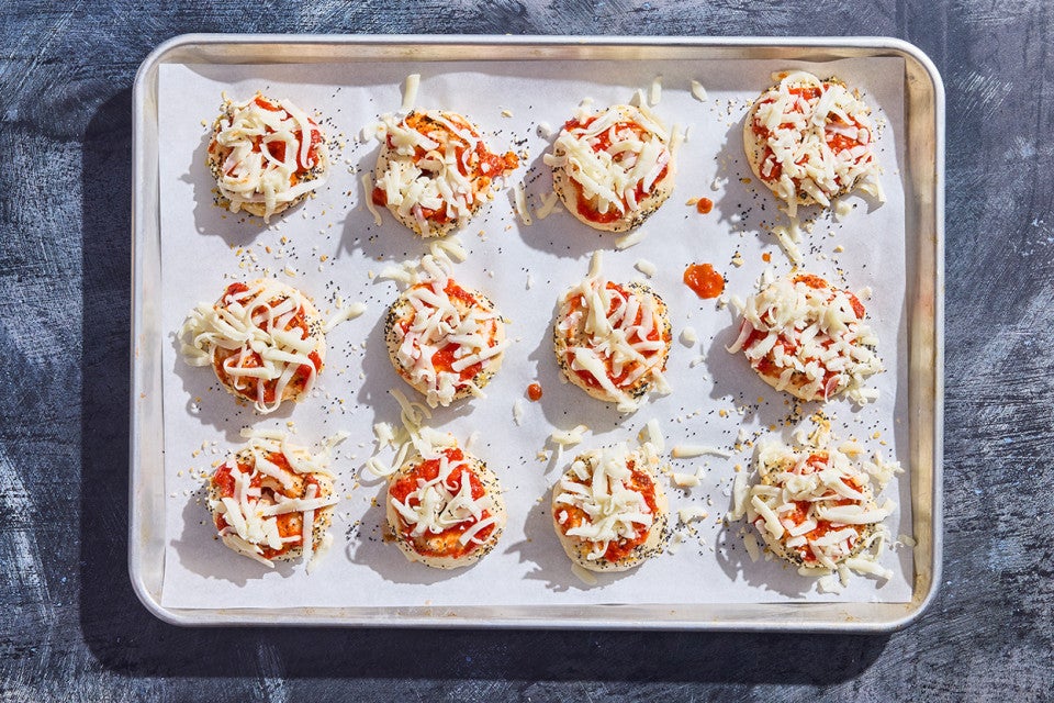 12 uncooked prepared mini pizza bagels on a sheet tray ready to go into the oven.