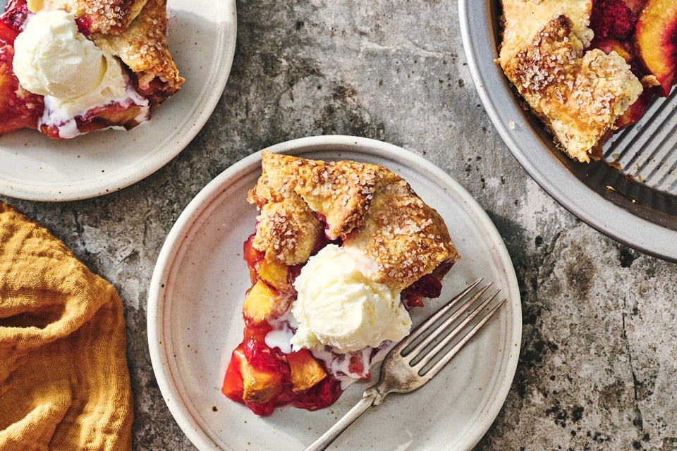 A slice of Rustic Raspberry-Peach Pie topped with vanilla ice cream on a white plate with a fork next to it.
