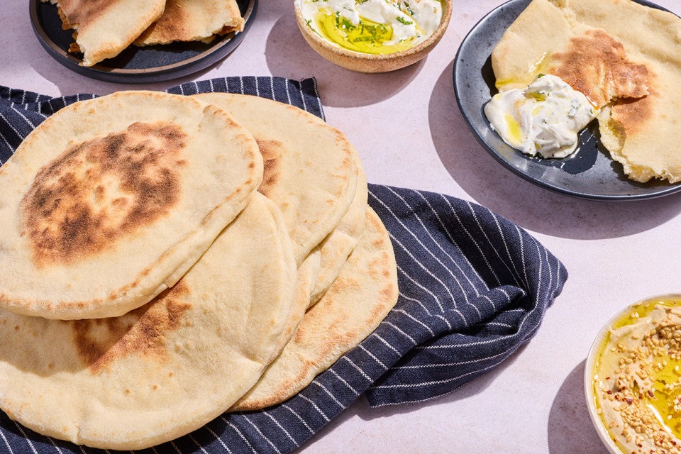 Finished Sourdough Pita Bread photographed from above in a stack on a dark blue napkin and dips and other bits spread around a table.