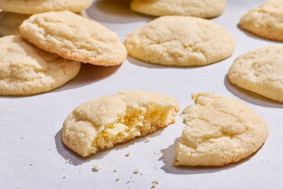 Sourdough Sugar Cookies laid out on a counter showing one of the cookies split in half with a slightly cakey interior and crumbs.