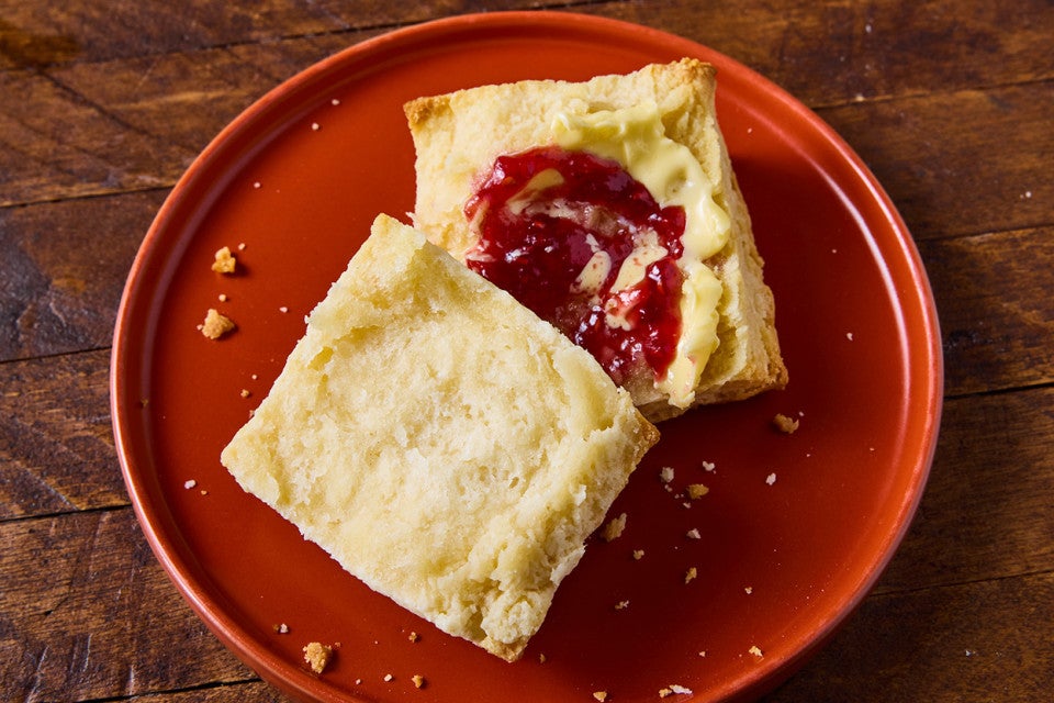 Overhead view of a sliced biscuit on a red plate and a wooden table. There red jelly and butter on the biscuit slice.