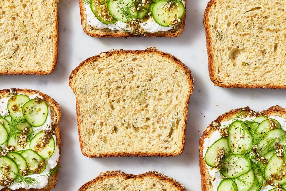 Multigrain Protein Bread slices arranged on a countertop and seen from above. Some have thin slices of cucumber on a spread while others are plain.
