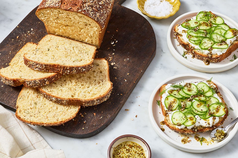 Multigrain Protein Bread on a dark brown cutting board and plates with slices that have cucumbers and spread on them.