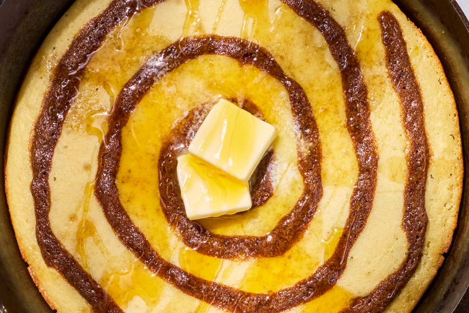 A swirled Cinnamon Roll Skillet Pancake in a skillet with dollops of butter on top and drizzled with maple syrup.