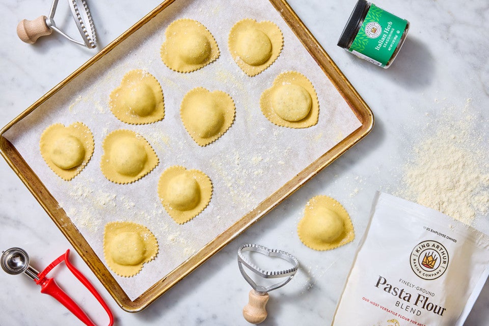 Homemade Cheese Ravioli shown cut into heart shapes and organized on a sheet tray covered in parchment with ingredients and tools strewn about a light marble counter.