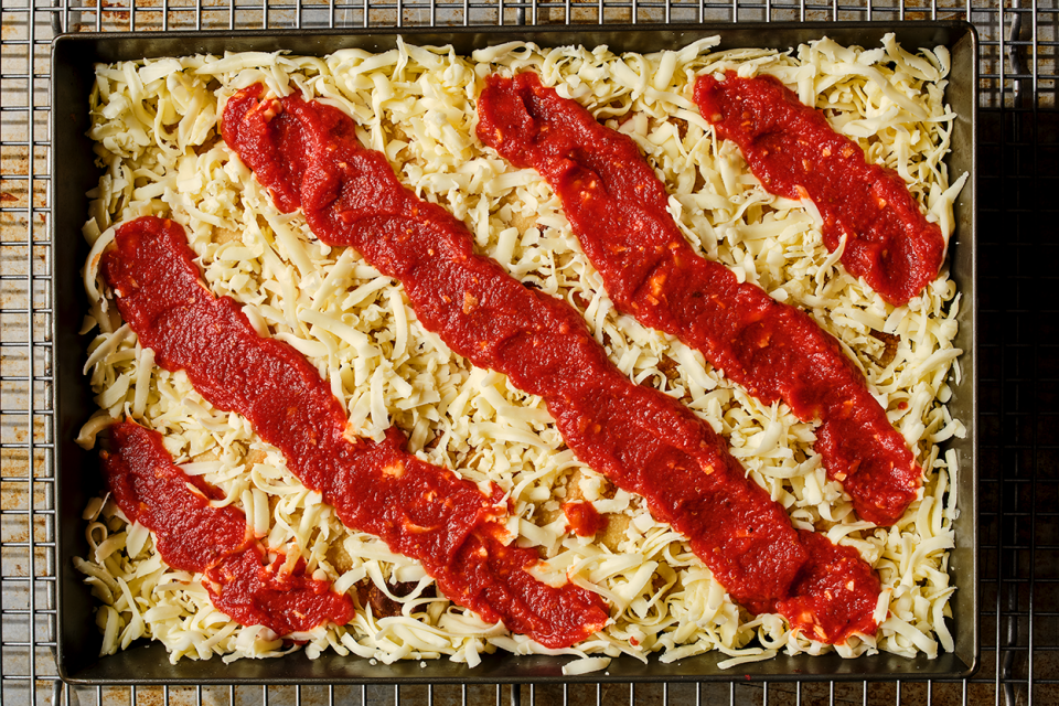 Preparing Detroit style pizza in a classic dark pizza pan with strips of sauce over the top of cheese.