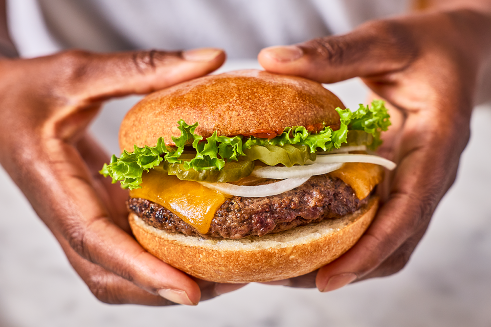 Holding a hamburger that is loaded up with cheese, onion, and lettuce all between a gluten-free roll.