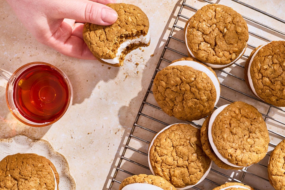 A hand holding an oatmeal cream pie that has a bite taken out of it with others on a cooling rack.